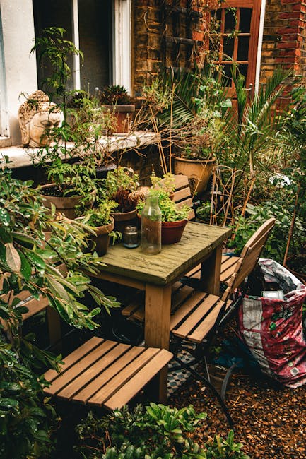 A cozy outdoor garden setting featuring a rustic wooden table surrounded by matching wooden and metal chairs, situated on a gravel surface. The table is decorated with various potted plants, including